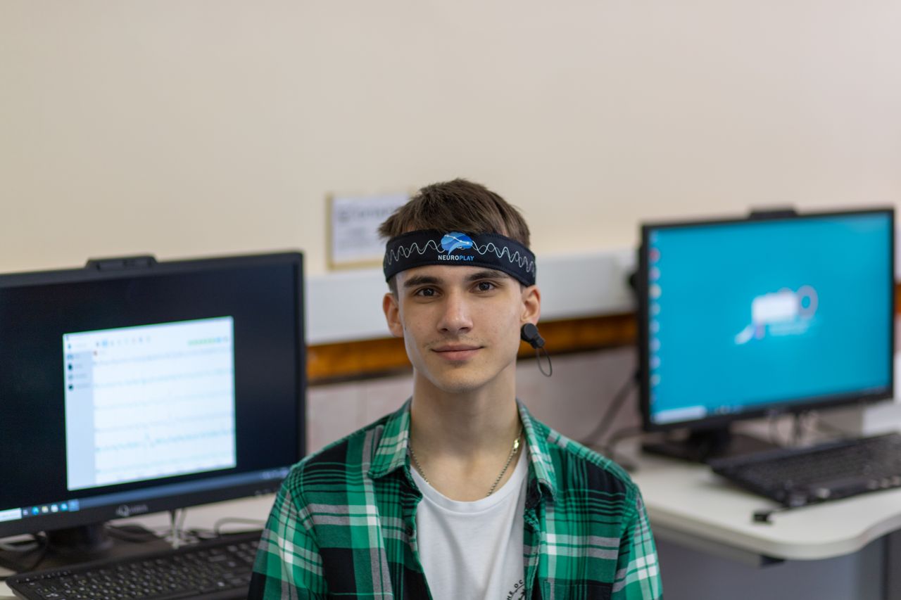 services-03 Portrait of a young man in a plaid shirt wearing a Neuroplay headband with computers in the background.