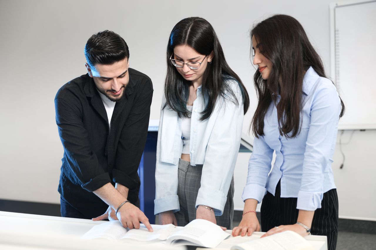 services-02 Three young adults discussing and pointing at open documents in a modern office, fostering teamwork.